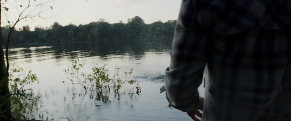 Man skips a stone across the river as water reflects woods on opposite shore