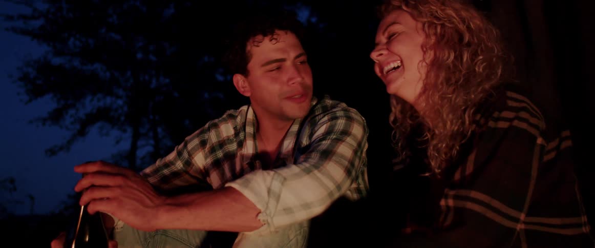 A man and woman sit outside by a fire and talk and laugh