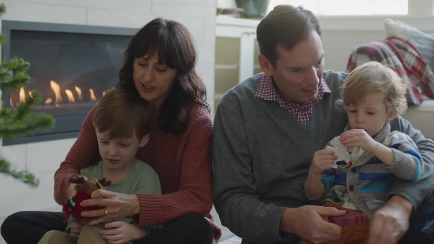 Man and woman each hold a young boy while sitting in front of a fireplace as the boys each play with a toy