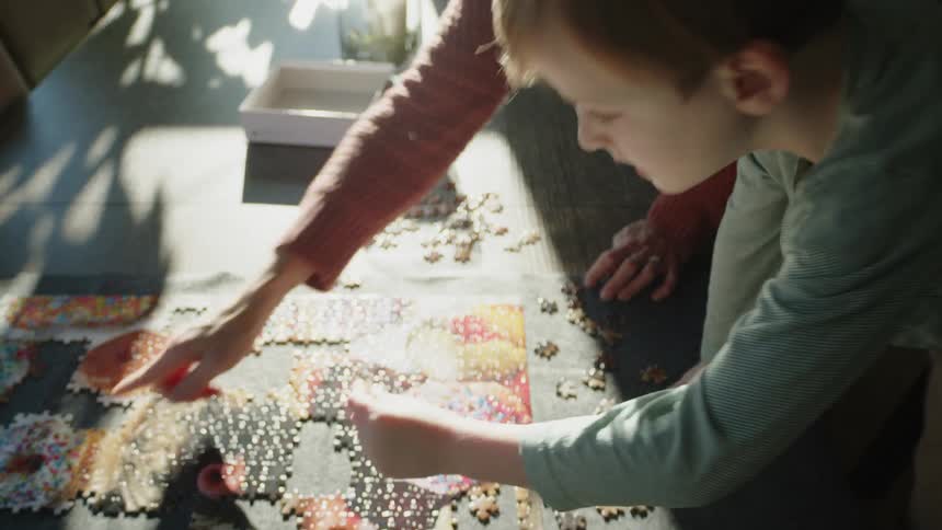 A woman points at a section of a puzzle on the table and a little boy stands and places a piece
