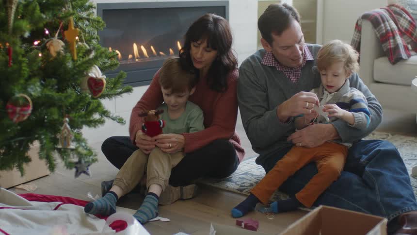 A man and woman each hold a child in their lap as the children play with toys while sitting on the floor in front of a christmas tree