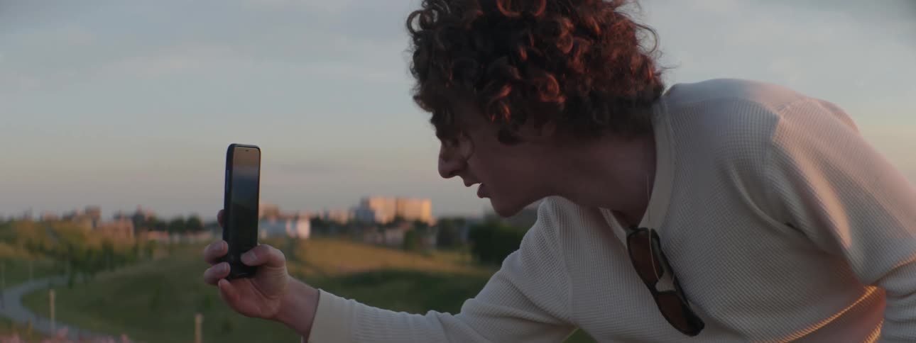 A man uses his smartphone to take a photograph of a hill covered in flowers on the outskirts of a city during the day