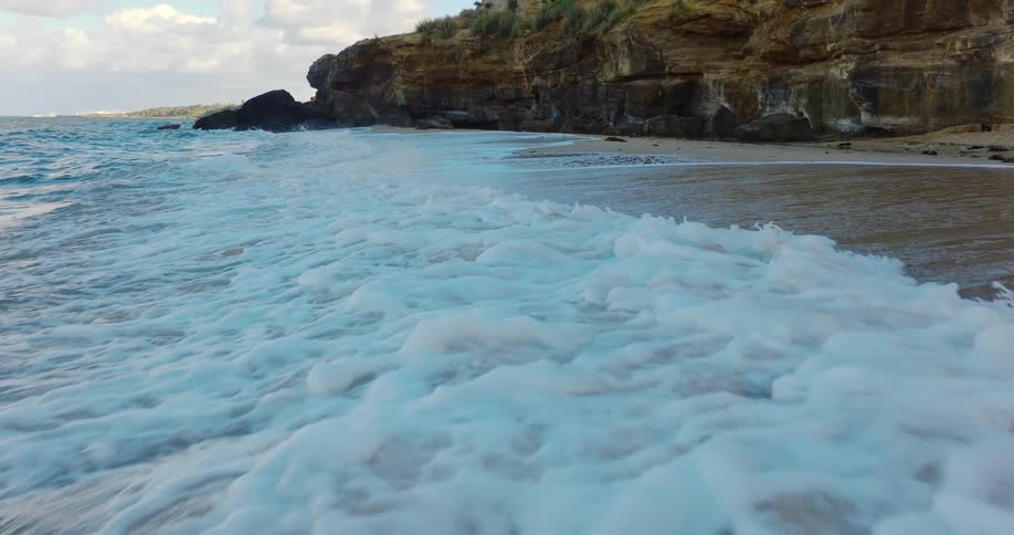 Numerous ocean waves crash upon a sandy shoreline near to some rocky cliffs