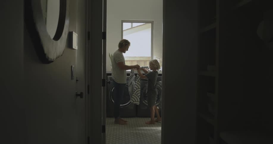 Father with wavy hair white shirt and pants shows son how to fold clothes in washing room