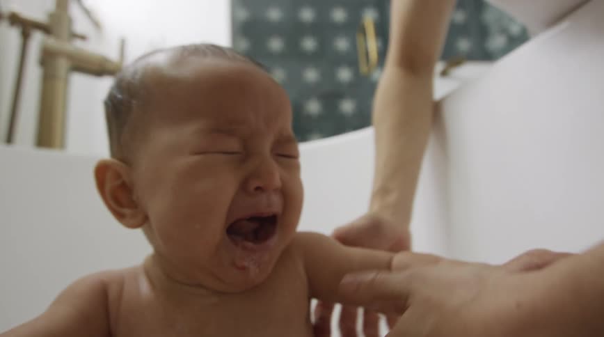 A baby sits in a bathtub and cries as his mother reaches for him