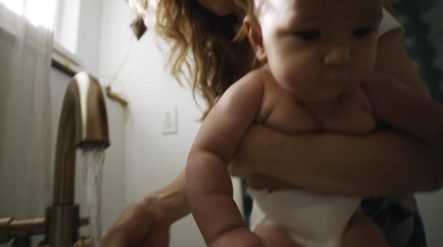 A woman holds her baby by a faucet and washes his hand