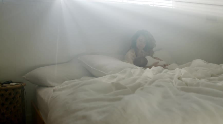 Mother with brown wavy hair sits on bed breastfeeding baby in living room at home