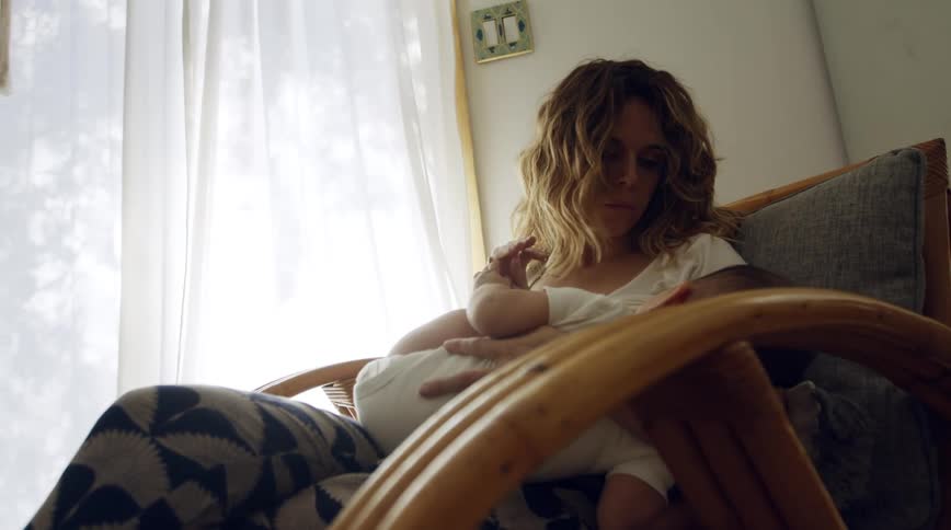 Mother with brown wavy hair and white shirt breastfeeds baby in armchair