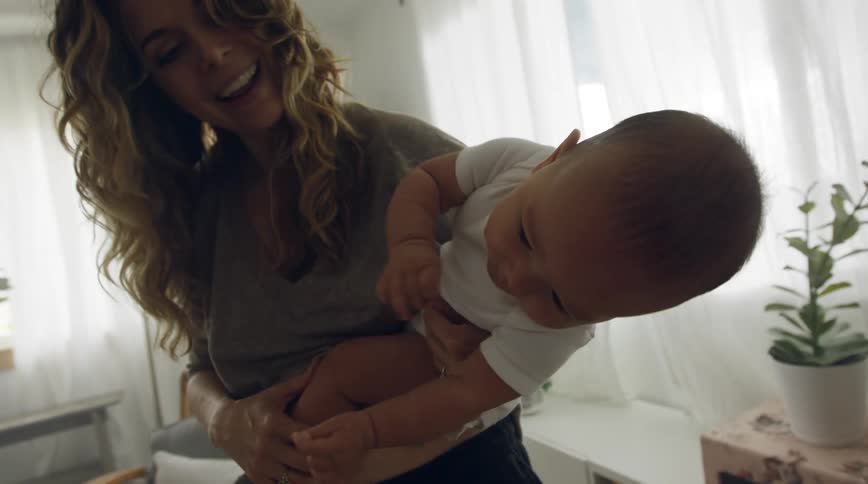 A woman laughs while carrying a baby indoors during the day