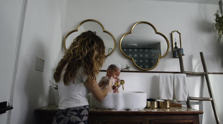 Mom gives baby a bubble bath in the bathroom sink basin