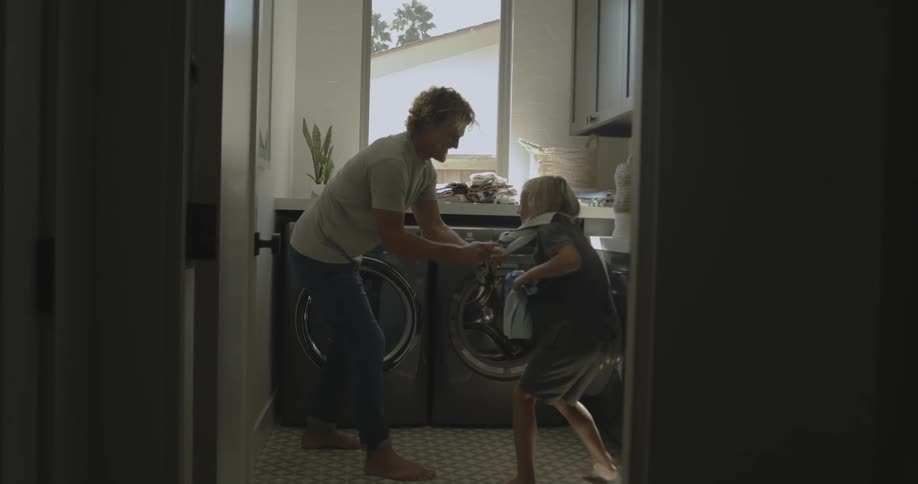 Father with wavy hair white shirt and jeans plays with son wearing t shirt and shorts in washing room