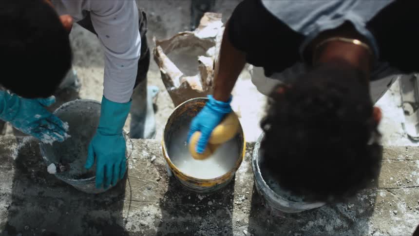 One man dips a sponge in a pail of water then squeezes the water out while a man next to him uses a tool and mortar