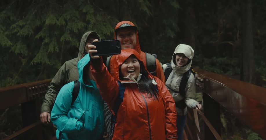 A woman takes a selfie with friends while standing on a pedestrian bridge in the woods
