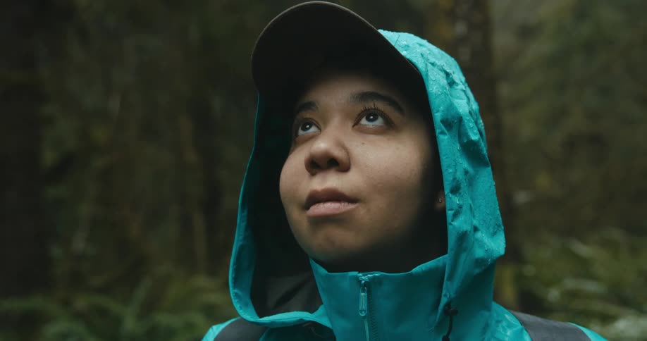 A woman in a raincoat looks up at tall trees in the forest