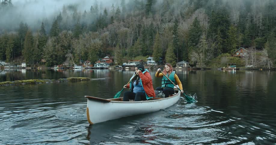 Two women row a canoe backwards in a lake toward shore