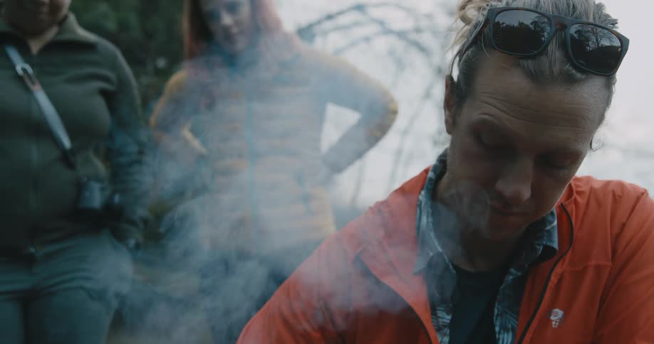 A man tends a fire outdoors as two women watch for nearby