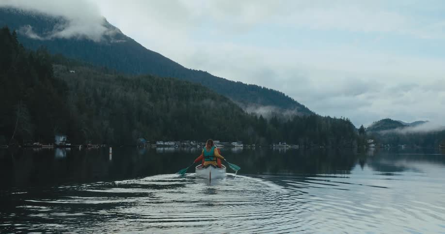 Two people uses oars to row a boat in a lake below mountains