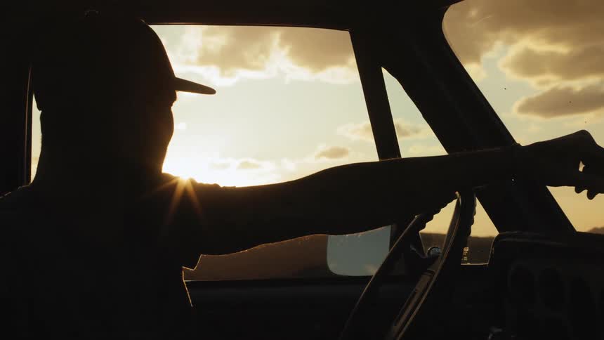 The silhouette of a man with his arm resting on a truck steering wheel looking at the glaring sun