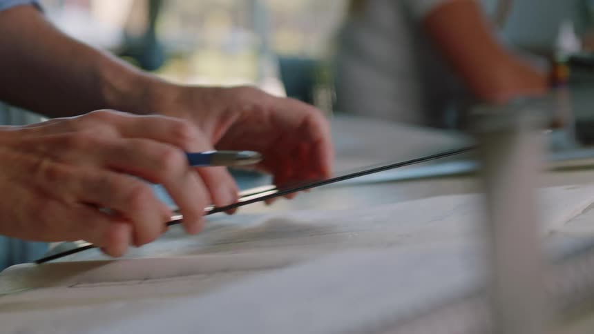 Someone is sitting at a desk with a pen in their hand and usinga plastic triangle to draw a straight line
