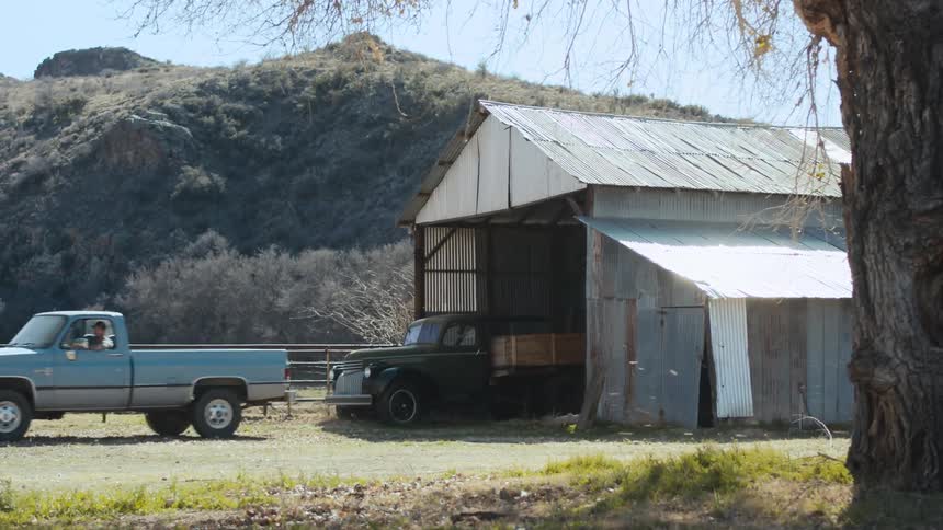 A man drives a truck from an aluminum shed in a rural area during the day