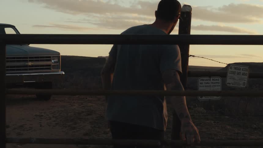 Tattooed man wearing a t shirt closes metal gate as truck sits in road nearby with mountains in the background under a red tinted horizon