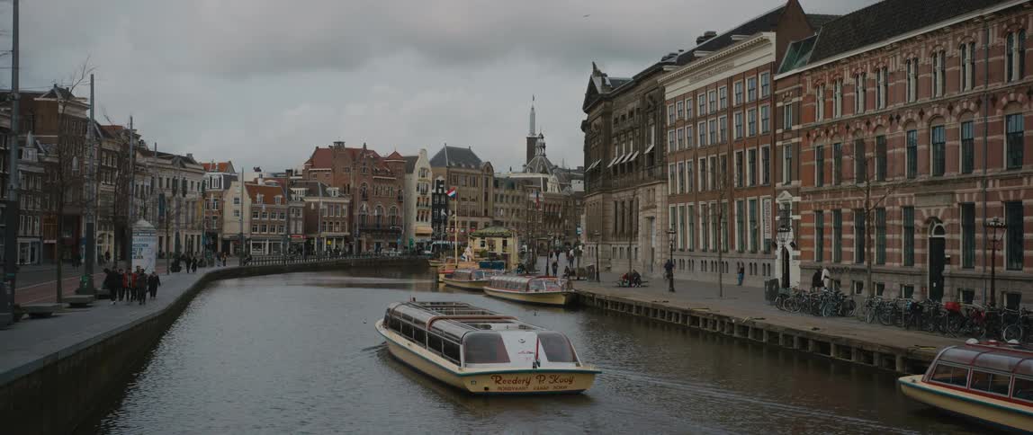 A tour boat travels a canal of Amsterdam alongside canal homes and streets
