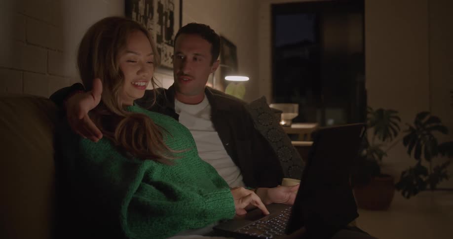 A woman uses a laptop while listening to a man sitting beside her on a sofa in the living room of a house at night