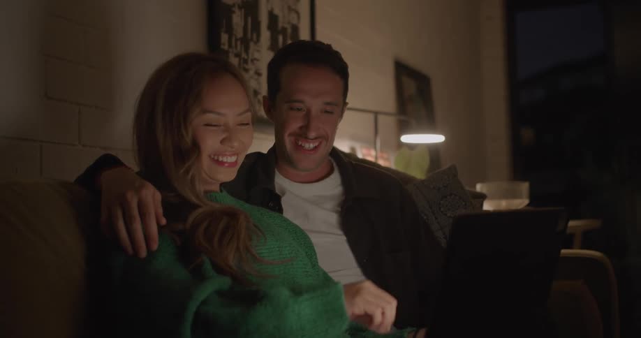 A woman points at the screen of her laptop while talking with a man sitting beside her on a sofa in the living room of a house at night