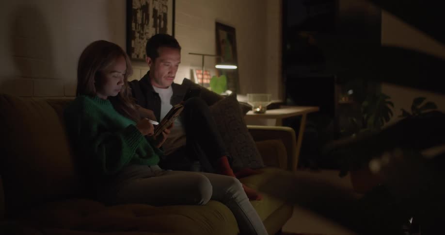 A woman and man silently focus on their smartphones while sitting beside one another on a sofa in the living room of a house at night