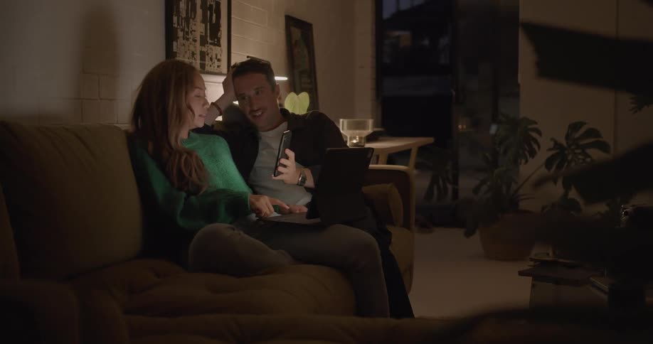 A man and woman sit on a couch together and the man looks at his cell phone and shows it to the woman and they laugh