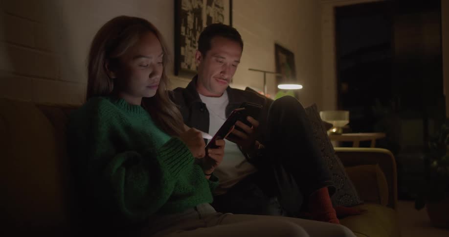 A man using his smartphone shows the screen to a woman using her own smartphone next to him on a sofa in the living room of a house at night