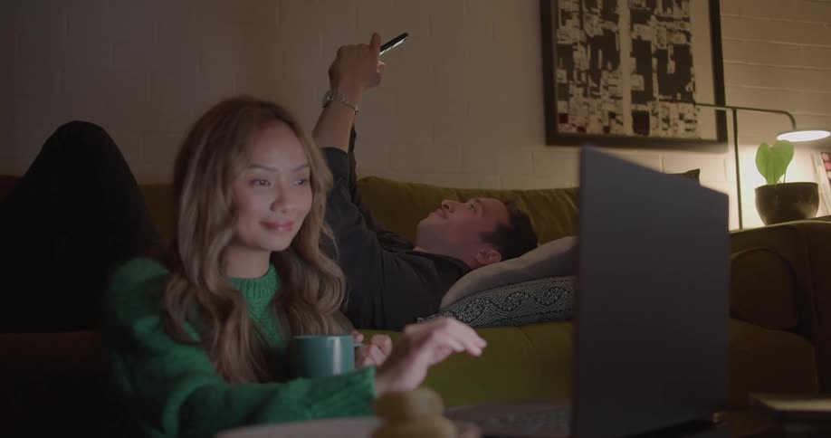 A woman sitting on the floor smiling and pointing using a laptop while her boyfriend lies on the couch using his phone