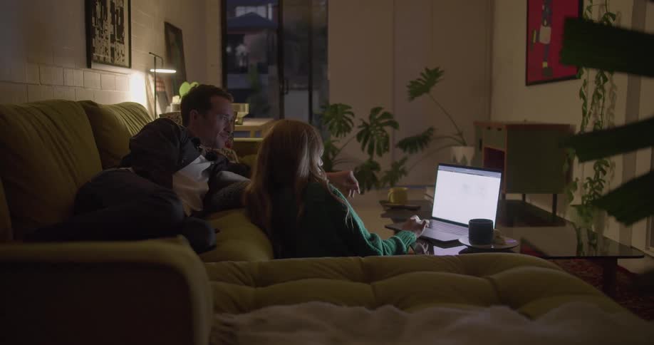 A woman sitting on the floor to use a laptop on a short table listens as a man lying on the sofa behind her speaks and points at the screen