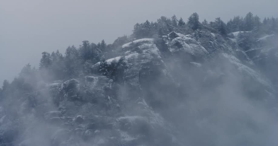 Snow dusted mountain and trees under a gray sky with fog swirling