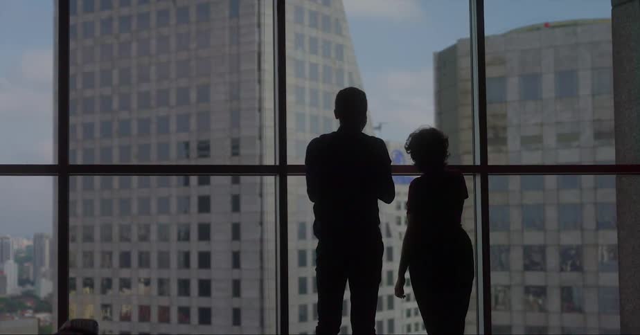 A man standing with a woman in front of a large window in a skyscraper uses his smartphone to photograph nearby buildings
