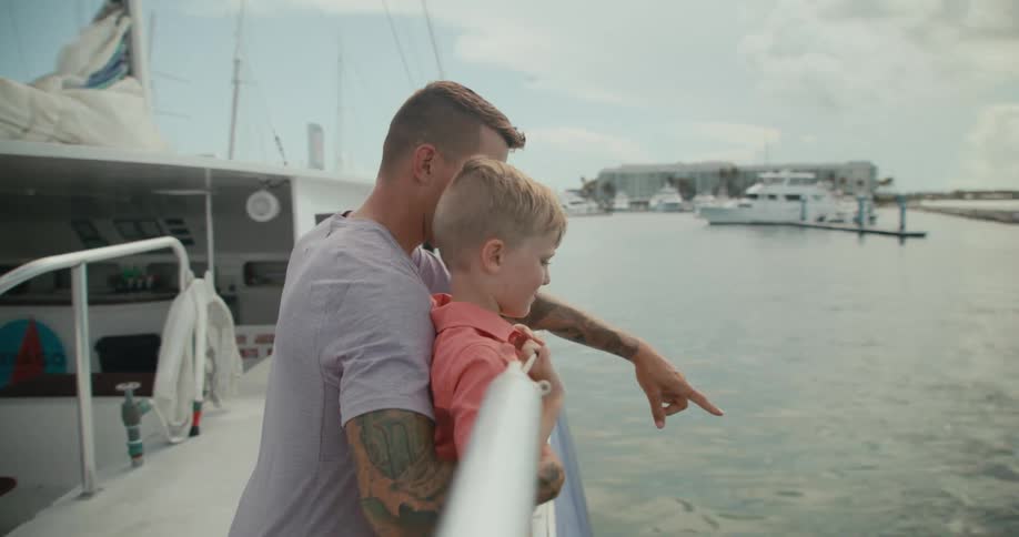 A man holds a boy standing near the railing on a boat and points at something in the water