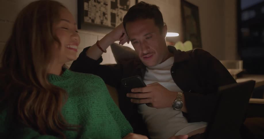 A woman points out something on the screen of a laptop to a man using his smartphone next to her on a sofa in the living room of a house at night