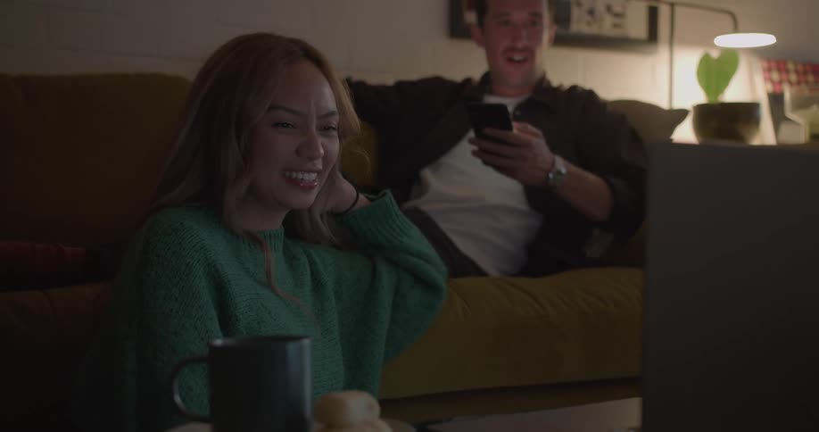 A man scrolls through his smartphone while also watching video on a laptop with his girlfriend