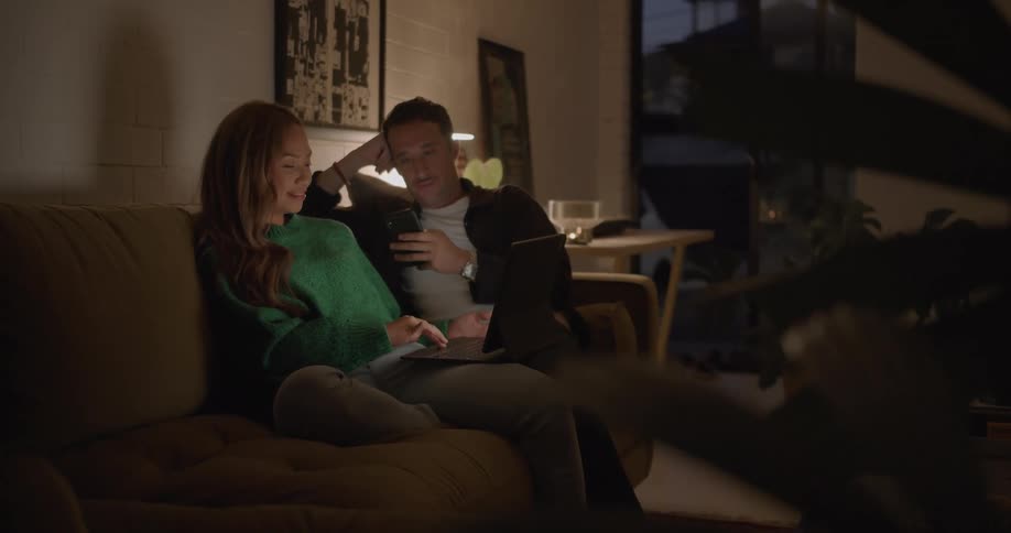 Man with mustache white and black shirt sits on brown couch next to woman with brown hair and green shirt using tablet in living room at home