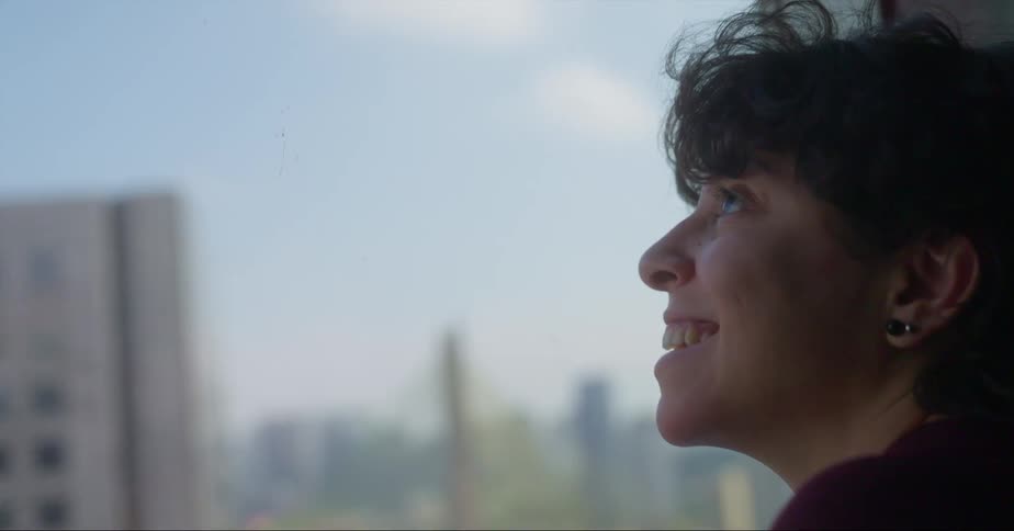 A woman standing near a window smiles as she looks out over an urban city skyline