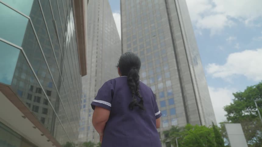 A woman stands at the base of a high rise office building in a city and looks up at it