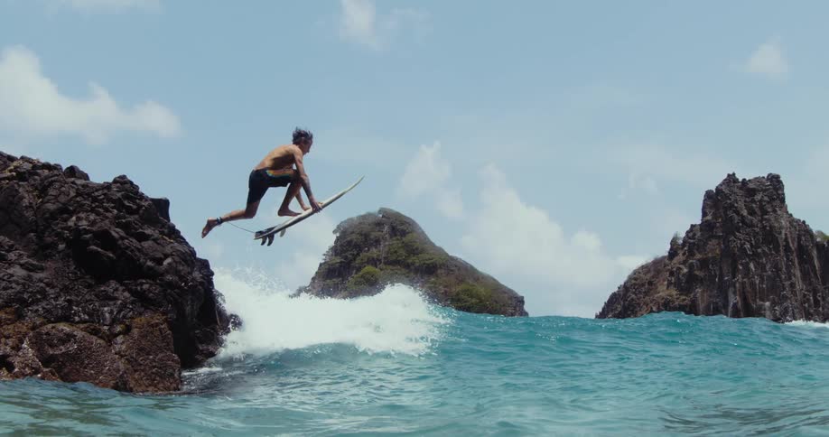 A male surfer leaps off of a rock formation and lands on his surfboard atop an ocean wave