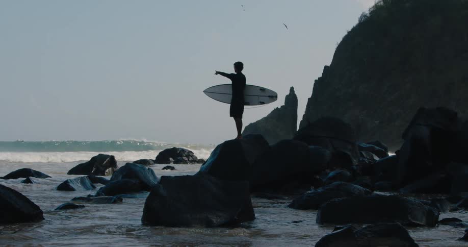 A male surfer holding his board stands on some rocks while pointing out to the ocean