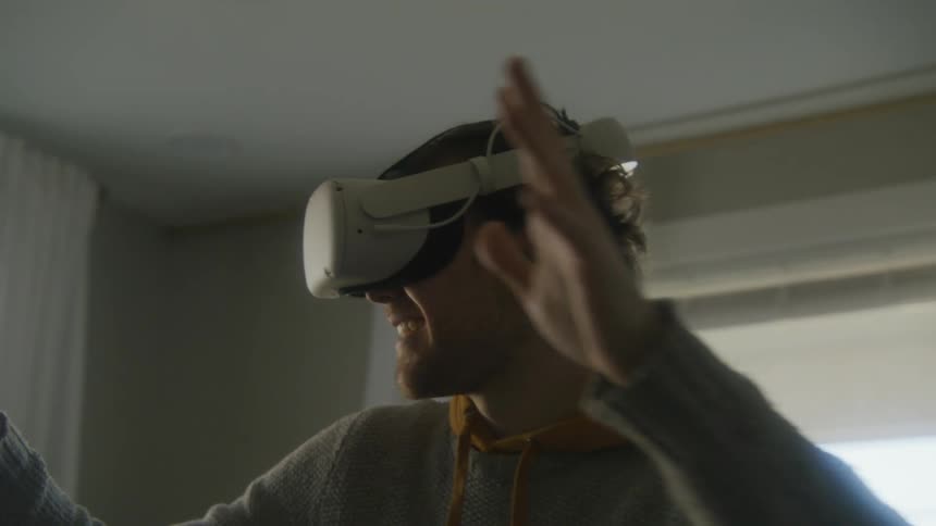 A teenage boy wears a virtual reality headset and turns in a circle and smiles at home