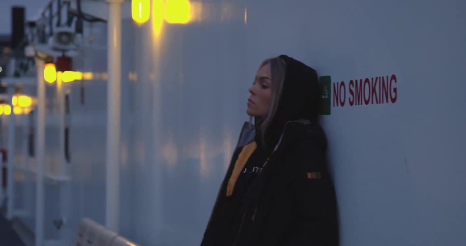 Woman wearing hooded jacket leans against wall near no smoking sign with city in the background