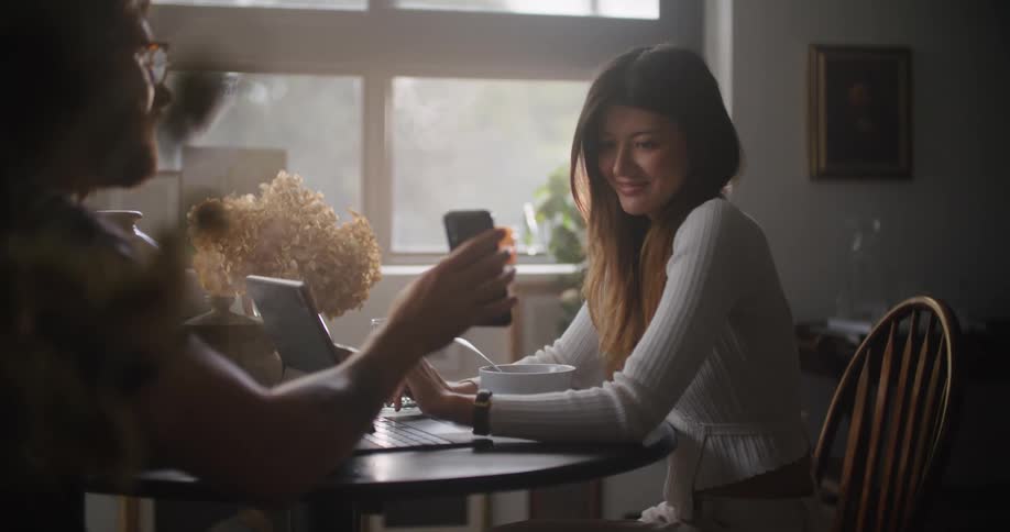 A man sitting at a table with a woman shows her his cell phone and she laughs and types on her laptop