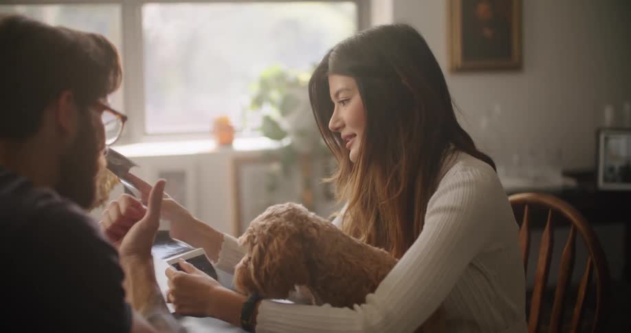 A man and woman sit at a table at home and look at ultrasound pictures while petting a dog that is in the womans lap