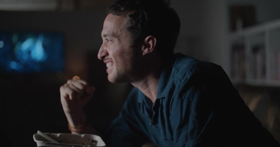 A man sits on a couch in front of his television while eating and pumping his fist in excitement