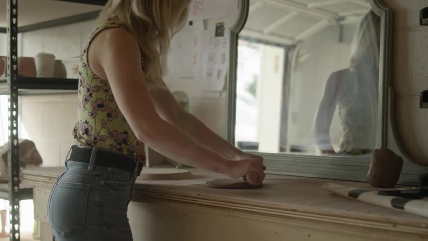 A woman with long blonde hair uses a rolling pin to flatten a piece of clay