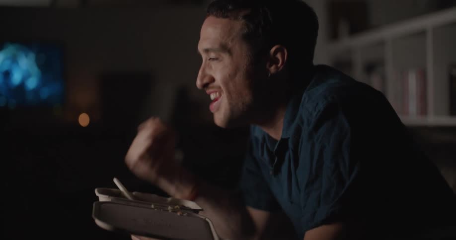 A man sits on a couch in front of his television while eating and pumping his fist in excitement